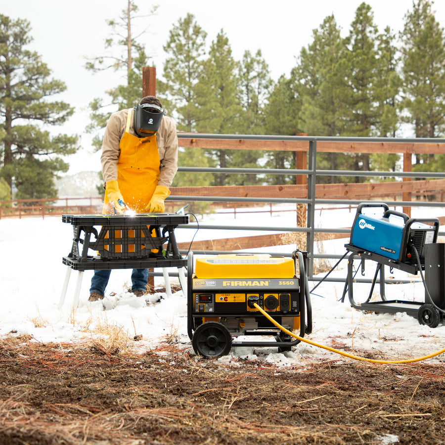 Dressed in outdoor workwear, a person uses tools at a portable table in a snowy yard, powered by a yellow FIRMAN Power Equipment 4450W Gas Portable Generator with CO alert, and another blue unit for backup—both connected with cables.