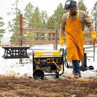 A man in a welding mask and protective leather apron works near a running FIRMAN Power Equipment Gas Portable Generator 7125W Recoil Start 120/240V in a snowy outdoor setting.