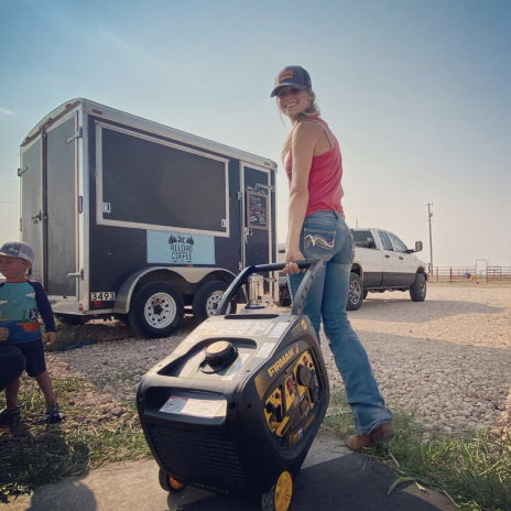 Image of a Woman holding a handle to an Inverter Generator and walking towards a pick up truck and trailer cart.