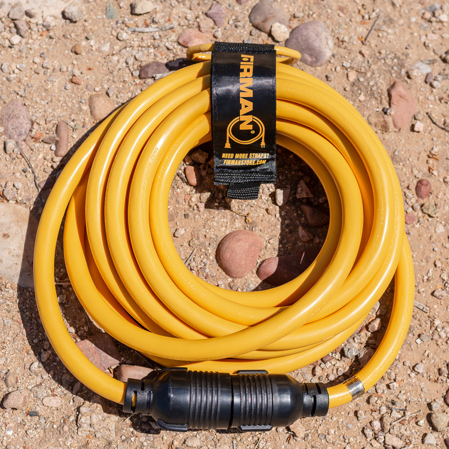 Coiled yellow FIRMAN Power Equipment 25' Heavy Duty L14-30P to L14-30R Power Cord Extension With Storage Strap on a gravel surface with visible label and connected ends.