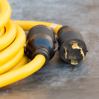 A close-up of a yellow FIRMAN Power Equipment 25' Heavy Duty L14-30P to L14-30R Power Cord Extension With Storage Strap, featuring a male plug and a female socket on a concrete surface.
