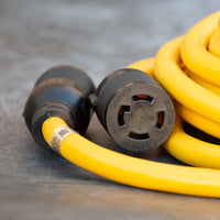 Close-up of a yellow FIRMAN Power Equipment 25' Heavy Duty L14-30P to L14-30R Power Cord Extension With Storage Strap, designed for outdoor use, set on a textured gray surface.