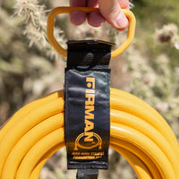 Close-up of a hand holding a yellow extension cord with a FIRMAN Power Equipment Heavy Duty Storage Strap With Handle labeled "FIRMAN" in a natural setting, with a cactus blurred in the background.