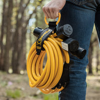 Someone holds the FIRMAN Power Equipment 25' Heavy Duty TT-30P to (3)5-20R Portable Generator Power Cord with black connectors and a storage strap while standing outdoors among trees.