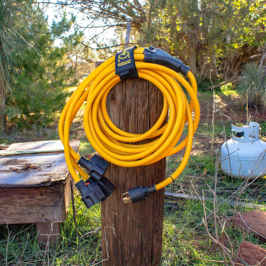 A 25' FIRMAN Power Equipment Heavy Duty L5-30P to (3) 5-20R Power Cord with storage strap is coiled and hanging on a wooden post outdoors, near a propane tank and bench among trees and grass.