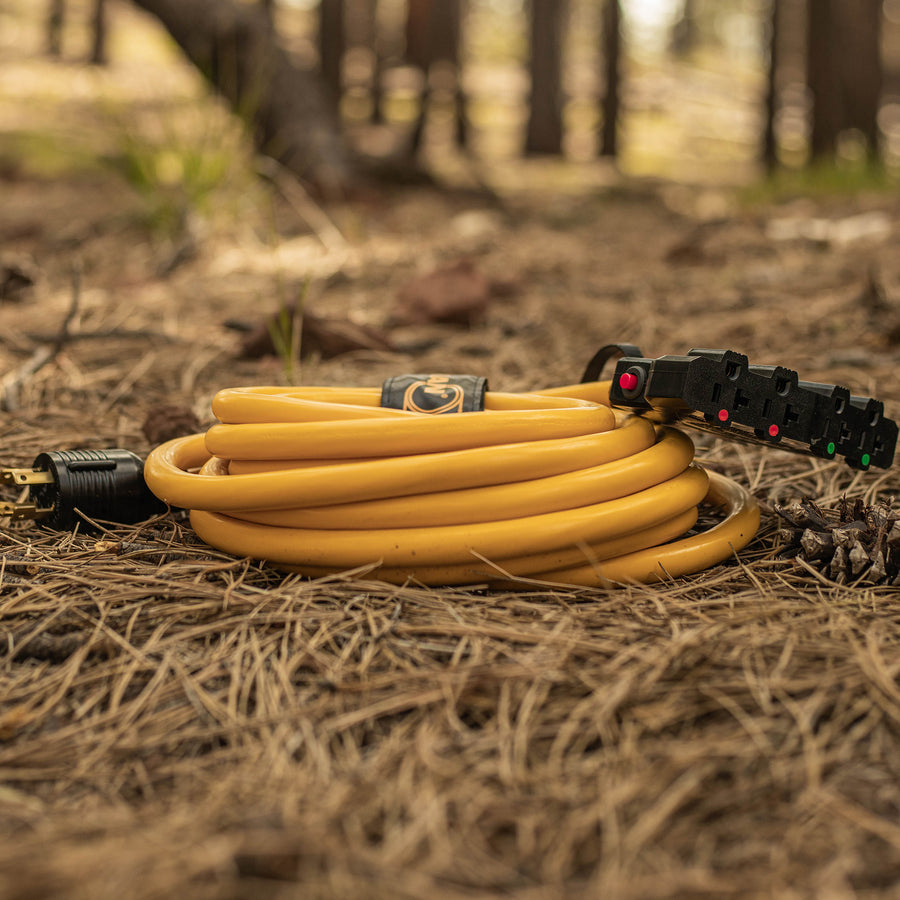 A coiled FIRMAN Power Equipment 25' Heavy Duty L14-30P to (4) 5-20R power cord with a black plug lies on a forest floor covered with pine needles.