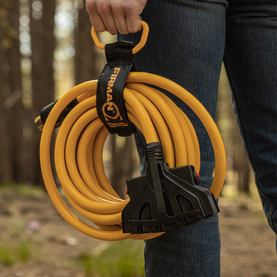 Outdoors, a person holds the FIRMAN Power Equipment 25ft Heavy Duty L14-30P to (4) 5-20R Power Cord with plastic ends and a storage strap for easy carrying.