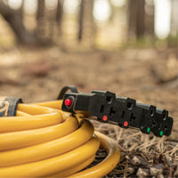 A FIRMAN Power Equipment 25ft Heavy Duty L14-30P to (4) 5-20R Power Cord with storage strap and black four-outlet adapter is coiled on the ground outdoors in a wooded area.
