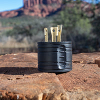 A portable black camping stove with extended metal fuel prongs, resting on a wooden surface against a backdrop of red rock formations and an FIRMAN Power Equipment L5-30P to TT-30R Adapter nearby.