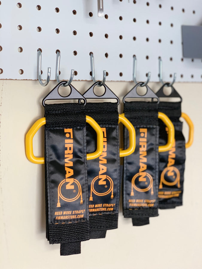 Black and yellow FIRMAN Power Equipment Heavy Duty Storage Strap With Handle hanging on hooks against a pegboard wall.