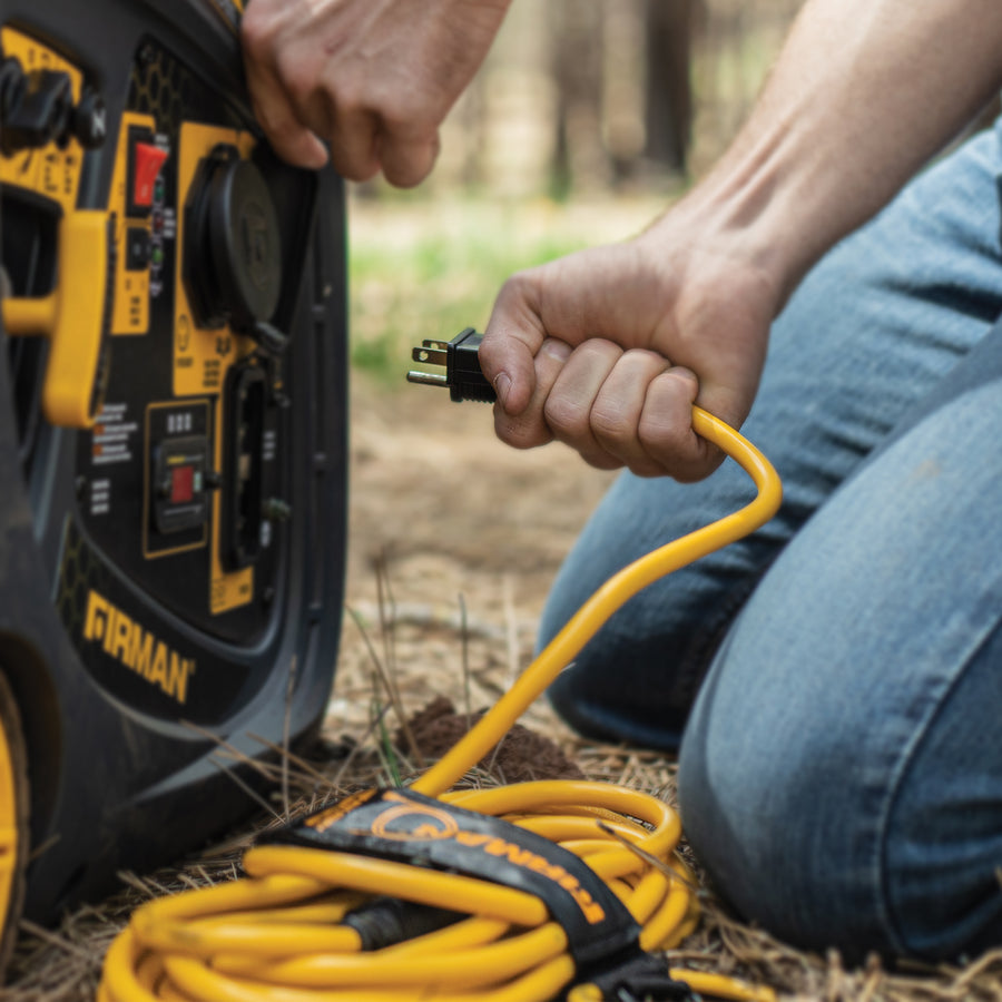 A person plugs a FIRMAN Power Equipment 25' Heavy Duty 5-15P to 5-15R Generator Utility Power Cord With Storage Strap into a portable generator in a forested area.