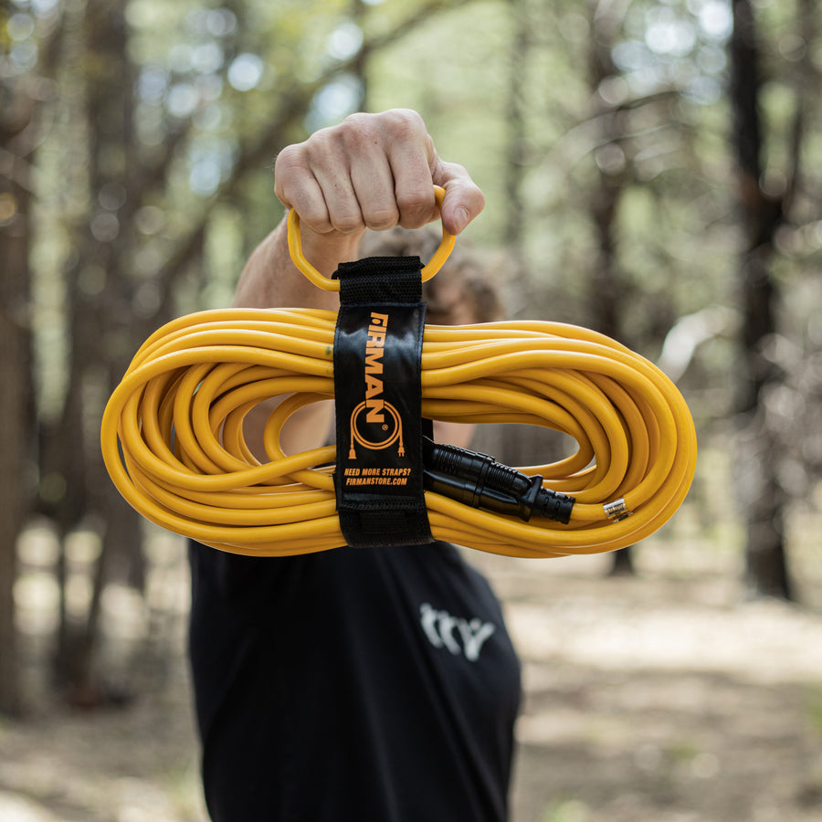 A person holds a 50ft Medium Duty 5-15P to 5-15R Generator Utility Power Cord with Storage Strap by FIRMAN Power Equipment outdoors in a wooded area.