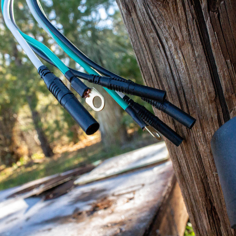 Close-up of electrical cables, including a FIRMAN Power Equipment 50A Parallel Cord Kit, hanging on a wooden post outdoors, with sunlit trees in the background.
