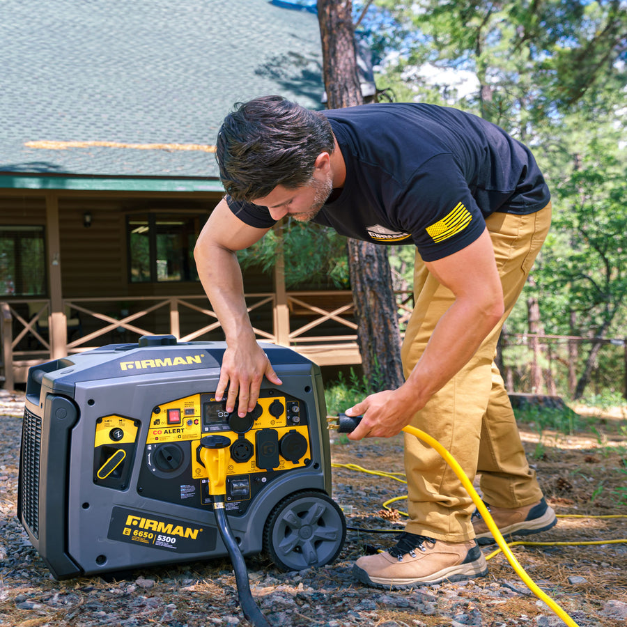 A man in a black shirt and tan pants plugs a yellow cord into a FIRMAN Power Equipment Gas Inverter Portable Generator 6650/5300 Watt 120/240V with CO Alert outside a house surrounded by trees.