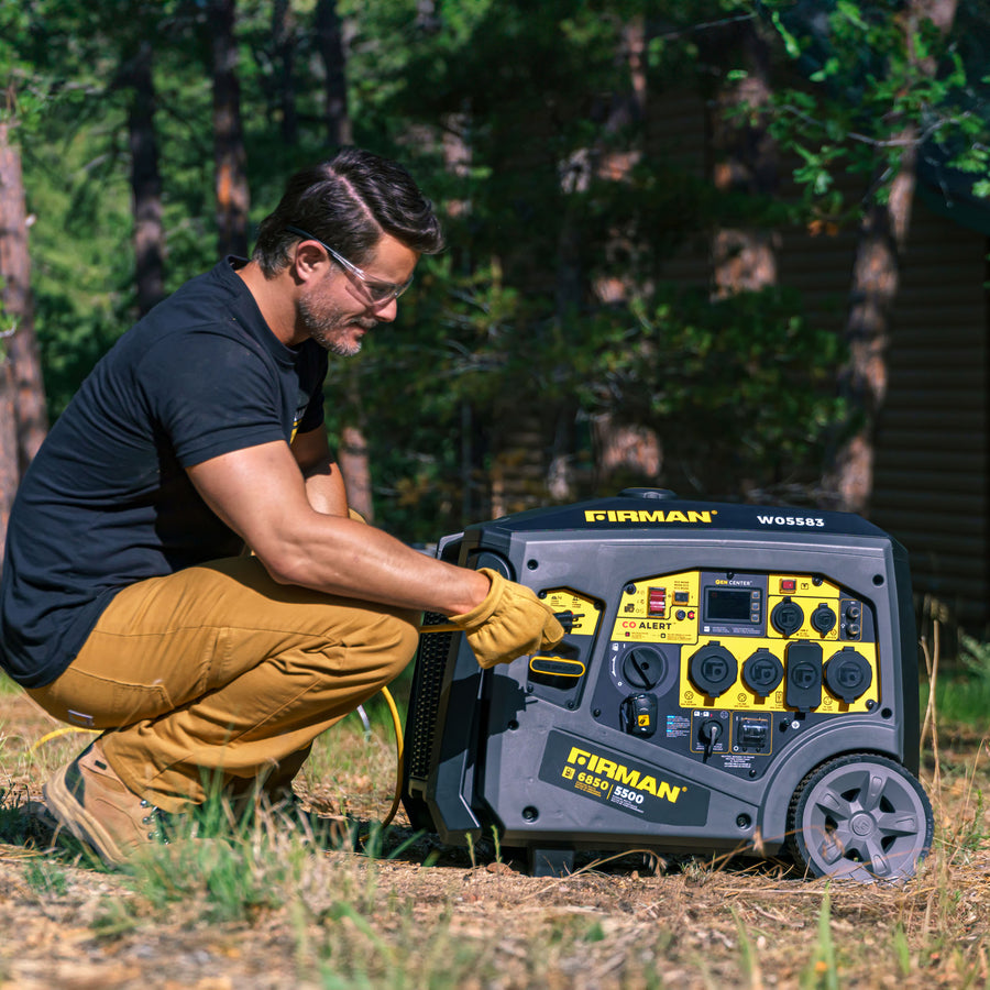 A man wearing safety glasses and gloves operates a FIRMAN Power Equipment Gas Inverter Portable Generator 6850/5500 Watt with Remote Start, featuring CO Alert, in eco mode outdoors near a wooded area.