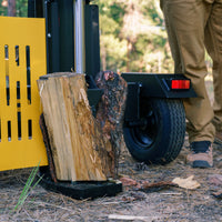 A close-up view of a log being split by the FIRMAN Power Equipment 27-Ton Log Splitter with a KOHLER engine. A person in brown pants stands nearby on a forest floor scattered with wood chips and pine needles.