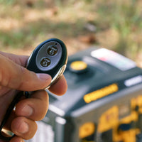 A hand holding a remote control with buttons labeled "Start" and "Stop." A yellow and black FIRMAN Power Equipment Gas Inverter Portable Generator 6850/5500 Watt Remote Start 120/240V CO Alert with an out-of-focus control panel is in the background.