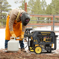 A person in protective gear connects a propane tank to a FIRMAN Power Equipment Dual Fuel 4550W Portable Generator Electric Start with CO Alert outdoors in a snowy, fenced area.