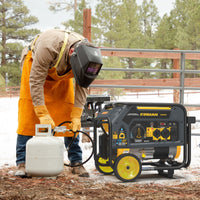 Wearing protective gear, a person connects a propane tank to the FIRMAN Dual Fuel Portable Generator 4550/3650W Recoil Start near a snowy fence, ensuring reliable winter energy with FIRMAN Power Equipment.