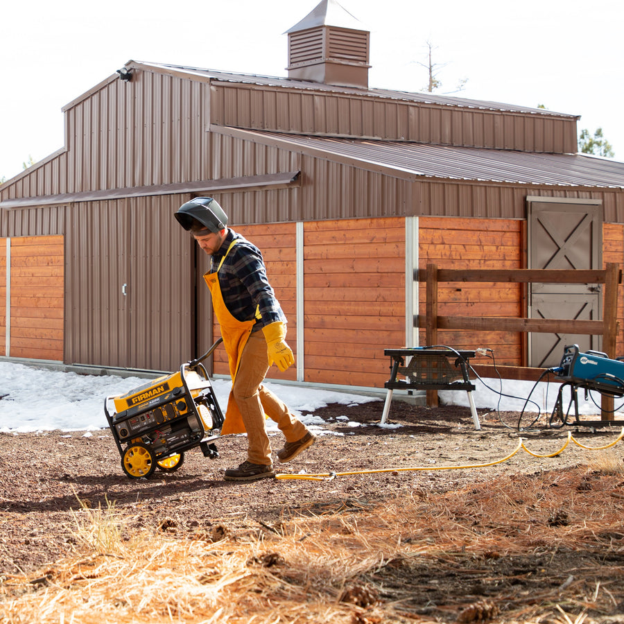 A person in a helmet and yellow overalls pulls a FIRMAN Power Equipment Gas Portable Generator 5000W Remote Start 120V towards a wooden barn on a snowy ground.