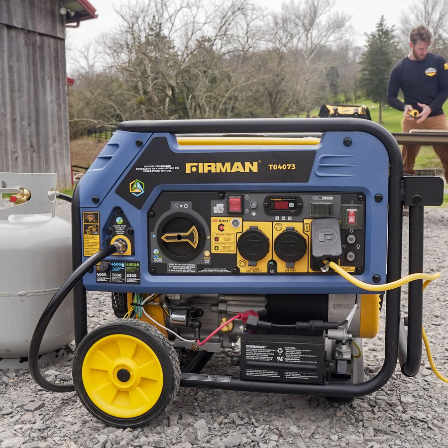 A blue FIRMAN Power Equipment Tri Fuel Portable Generator 4000W Electric Start 120/240V with CO ALERT on wheels, with various sockets and dials, operates outdoors while a man works in the background.