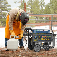A person in protective gear connects a propane tank to a FIRMAN Power Equipment Tri Fuel Portable Generator 7850/6300W Electric Start Gas CO Alert outdoors near a fence, with snow and trees in the background.