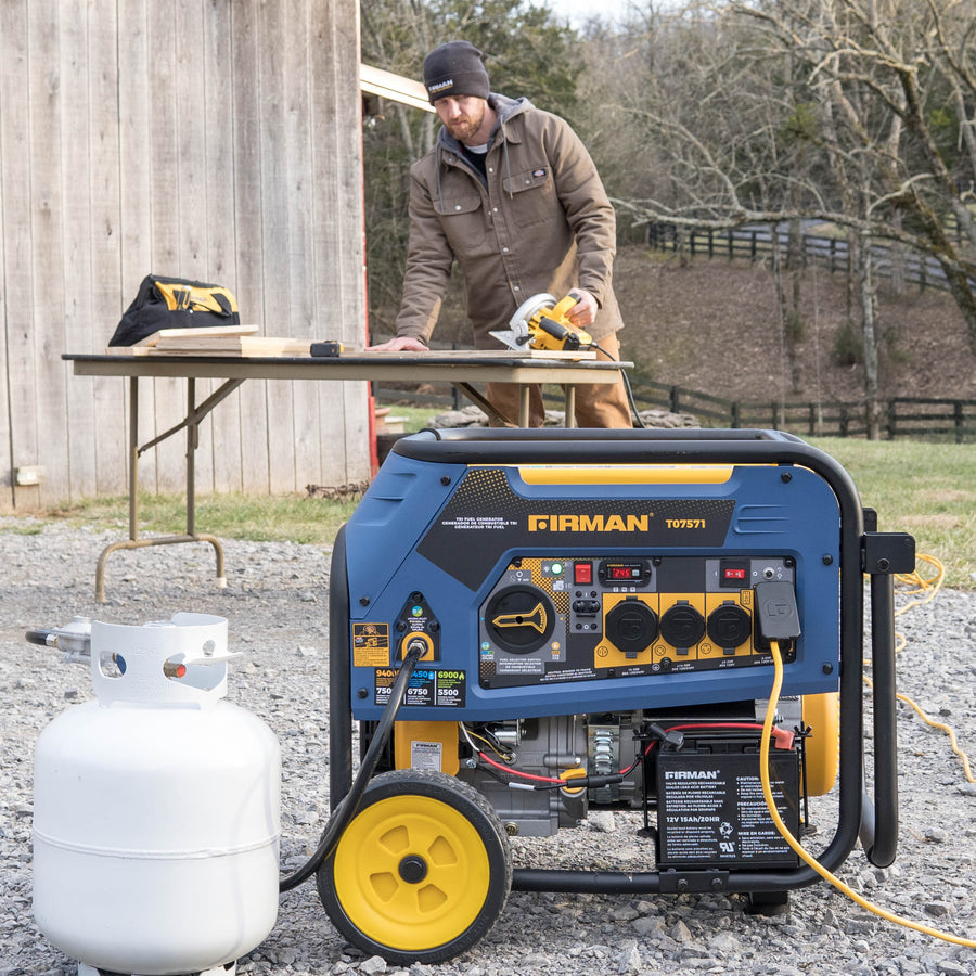 A FIRMAN Power Equipment Tri Fuel 7500W Portable Generator Electric Start 120/240V, connected to a propane tank, sits outdoors on gravel while a man works nearby—showcasing dependable home backup power.
