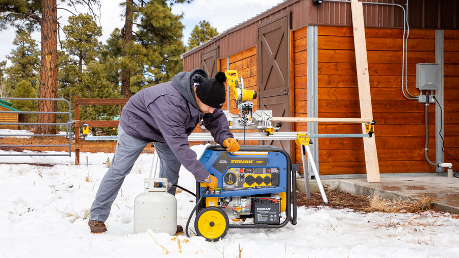 Man in winter clothing starting a FIRMAN Power Equipment Tri Fuel Portable Generator 8000W Electric Start 120/240V outside a wooden cabin in a snowy environment.