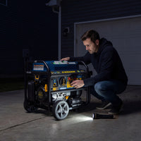 At night, a man kneels in the driveway using a FIRMAN Power Equipment Tri Fuel Portable Generator 8250/6600W Electric Start, with a flashlight beside him and the included wheel kit, cover, and cord nearby.