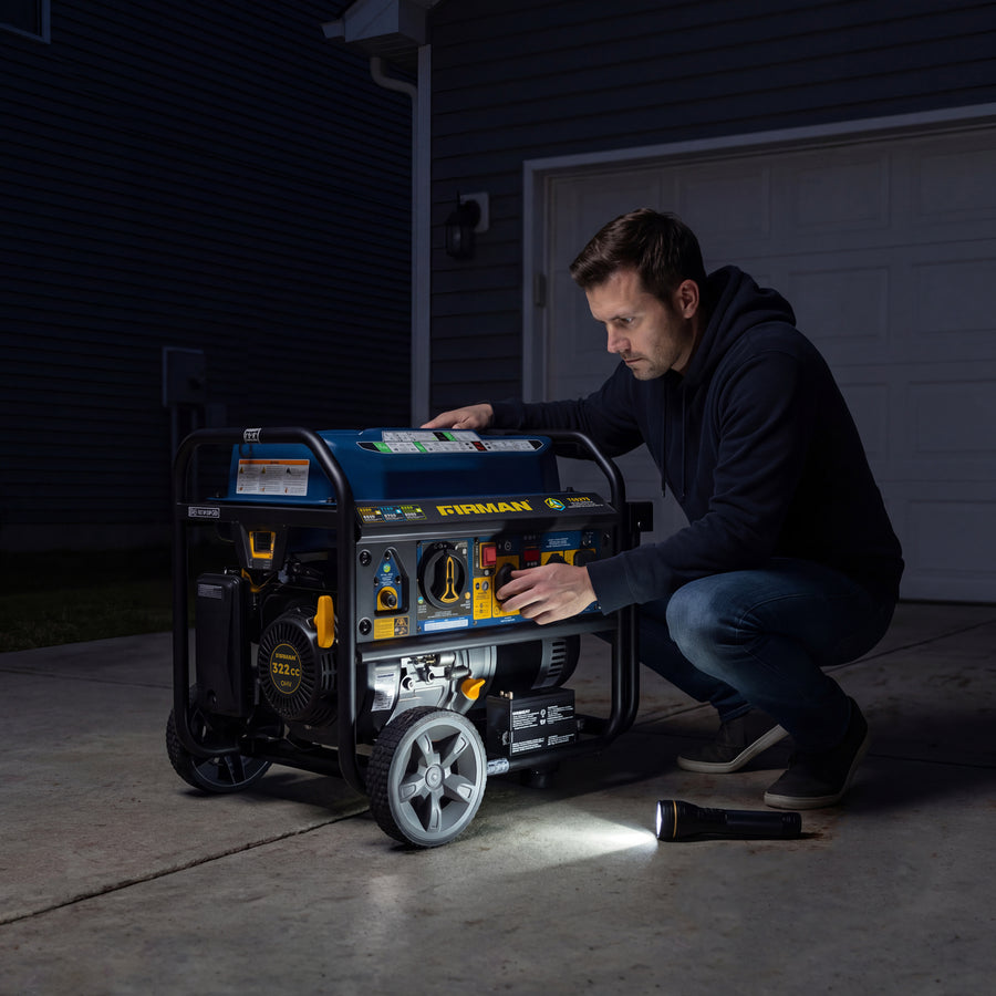 At night, a man kneels in the driveway using a FIRMAN Power Equipment Tri Fuel Portable Generator 8250/6600W Electric Start, with a flashlight beside him and the included wheel kit, cover, and cord nearby.