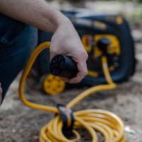 A man holding a black electrical plug with a yellow FIRMAN Power Equipment 25' Heavy Duty TT-30P to TT-30R Power Cord With Storage Strap, with a portable generator in the background.