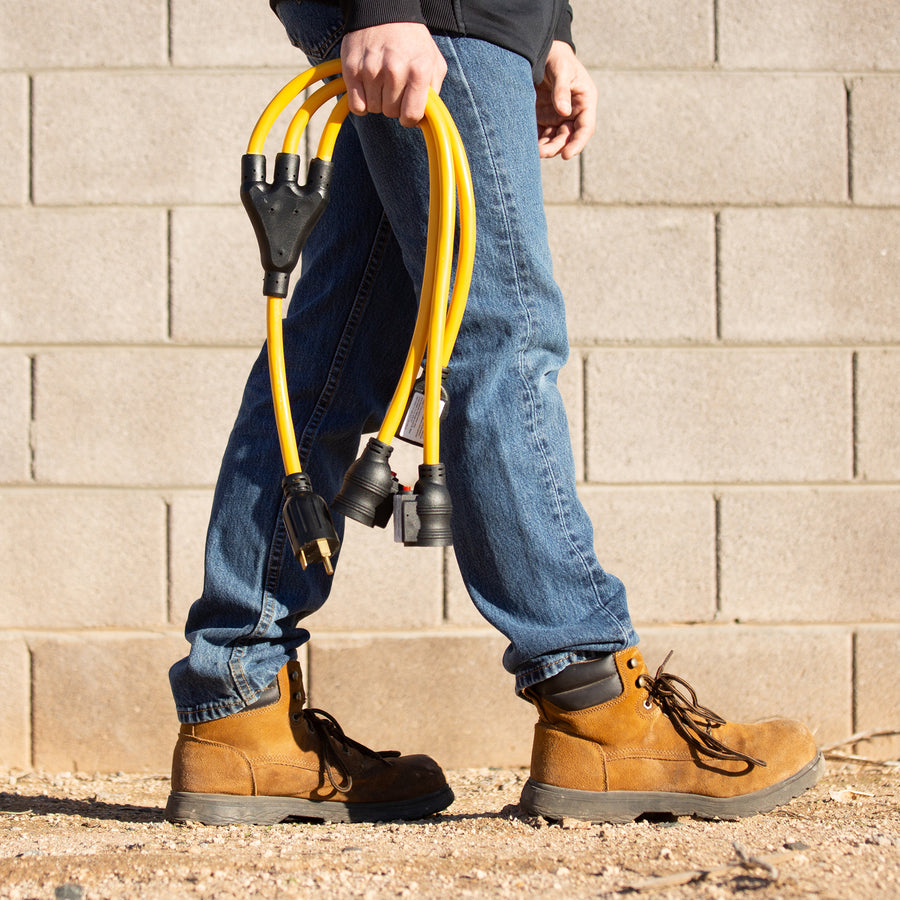 A person in jeans and work boots holding a yellow FIRMAN Power Equipment 3' Heavy Duty TT-30P to (3) 5-20R Short Power Cord, standing by a brick wall.