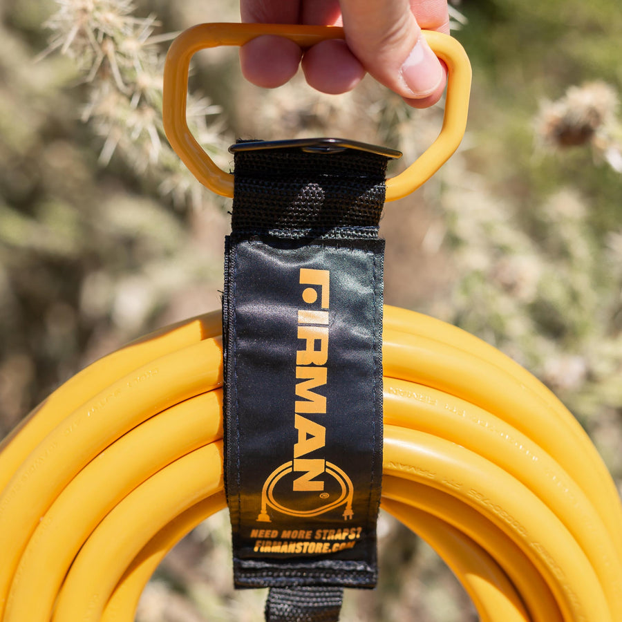 Close-up of a hand holding a yellow extension cord with a FIRMAN Power Equipment Heavy Duty Storage Strap With Handle labeled "FIRMAN" in a natural setting, with a cactus blurred in the background.