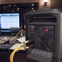 Back of a FIRMAN Power Equipment Zero E Portable Expandable Power Station with various cables connected, featuring expandable slide locking technology, set against a blurred background of a recording studio's equipment.