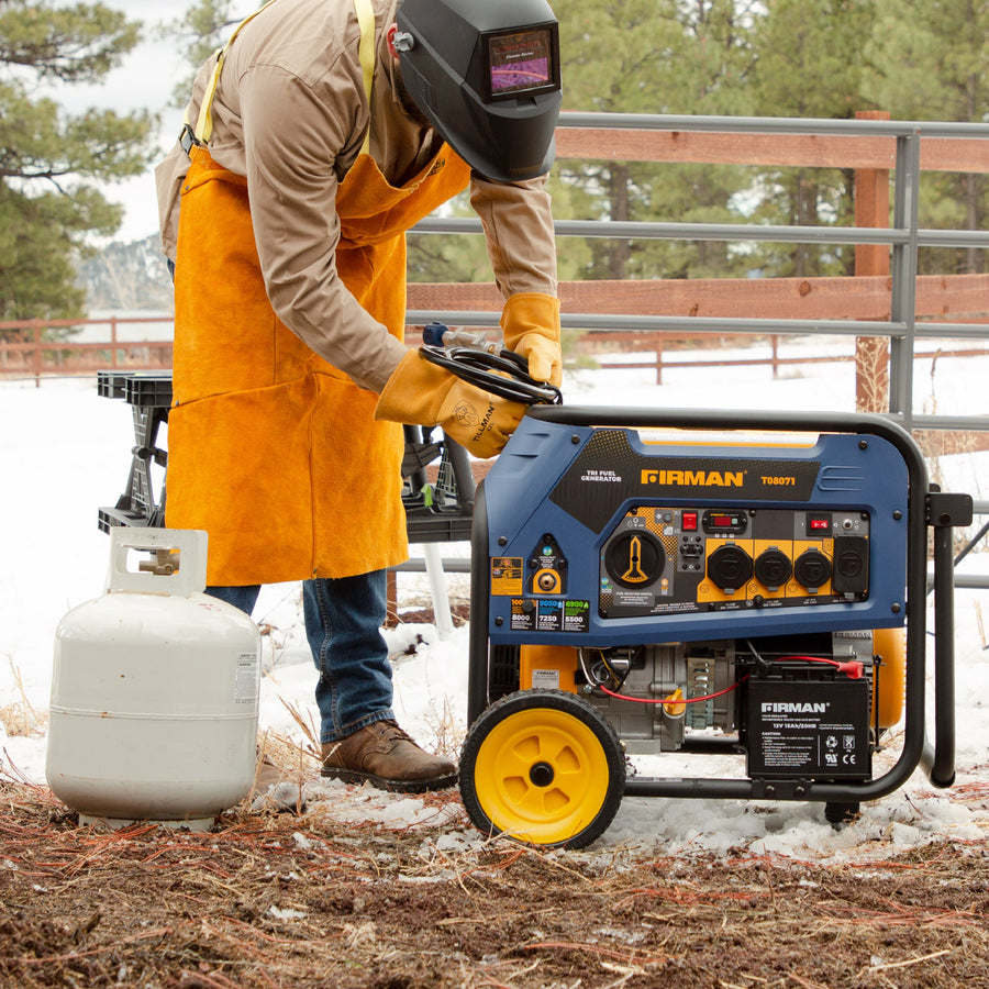 A person in a welding mask and apron adjusts a FIRMAN Power Equipment Tri Fuel Portable Generator 8000W Electric Start 120/240V next to a propane tank in a wooded area.