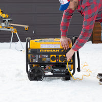 A person in a red plaid shirt starting a FIRMAN Power Equipment Gas Portable Generator 4550W Remote Start 120/240V for backup power in a snowy outdoor setting.